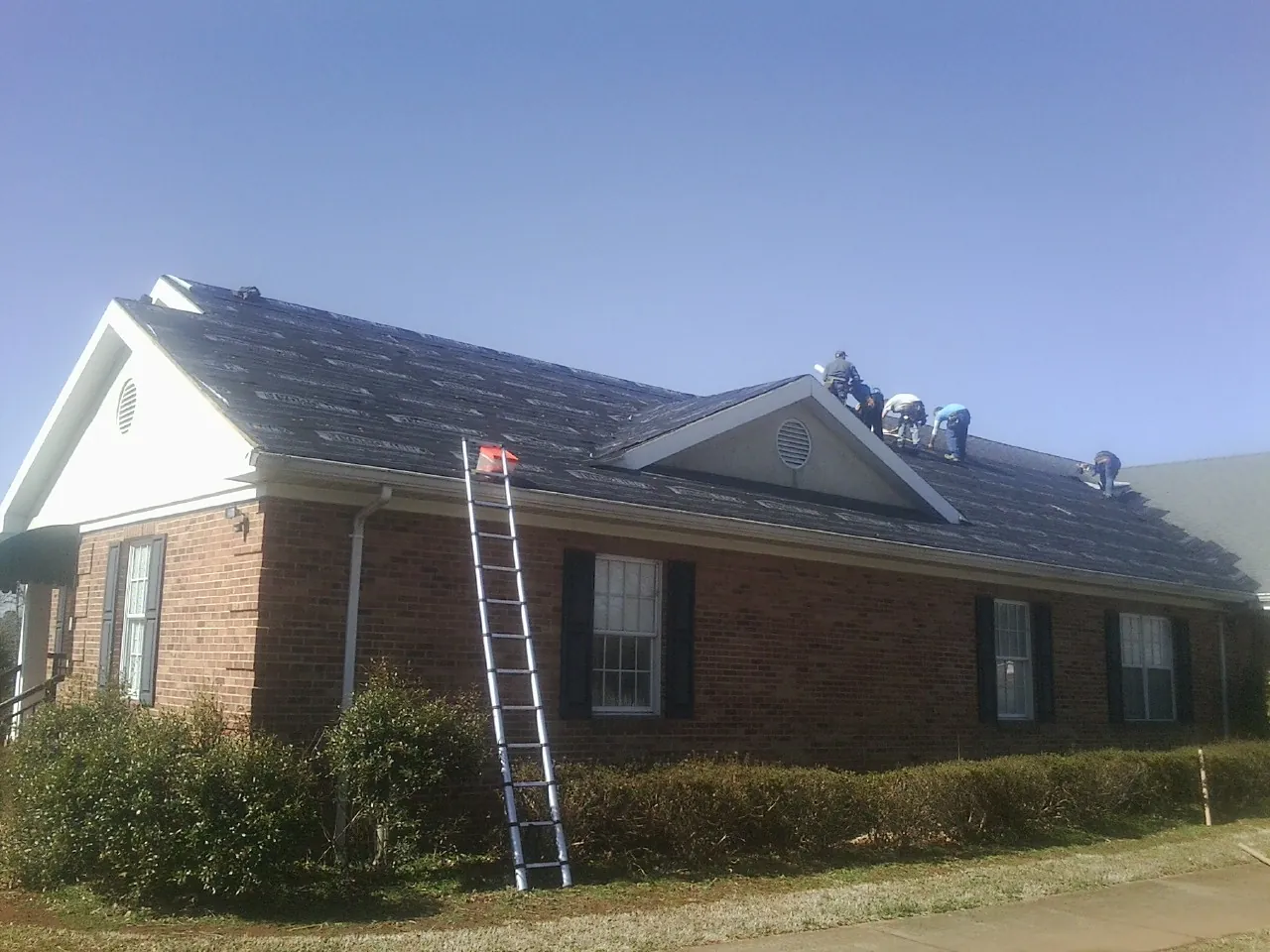 Crew tearing shingles off a brick ranch roof with a ladder set — Emergency Roof Repair work by our crew in Furman