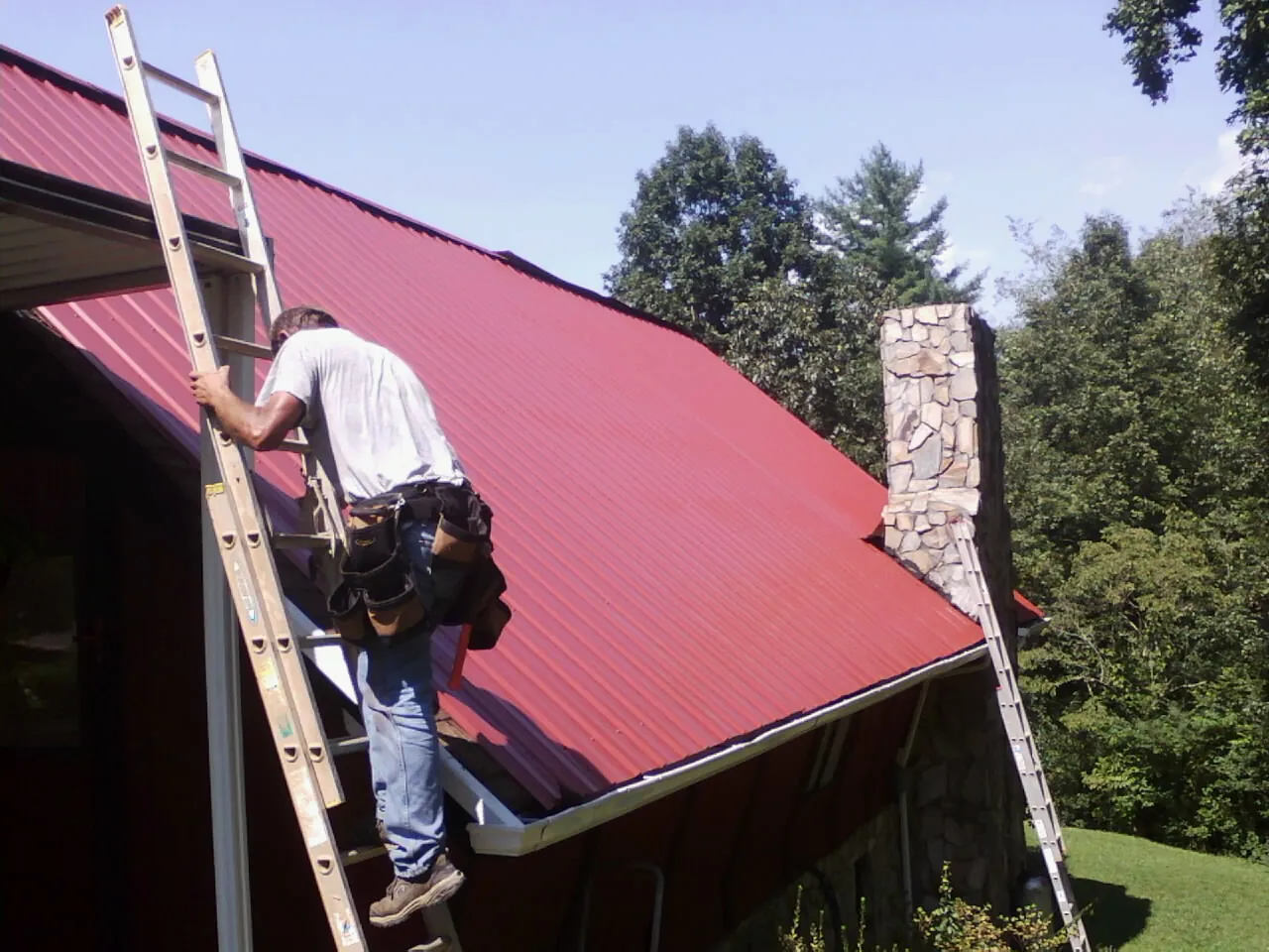 Roofer on a ladder beside a red metal roof and stone chimney — Soffit & Fascia Repair by Roofing Brothers in Furman